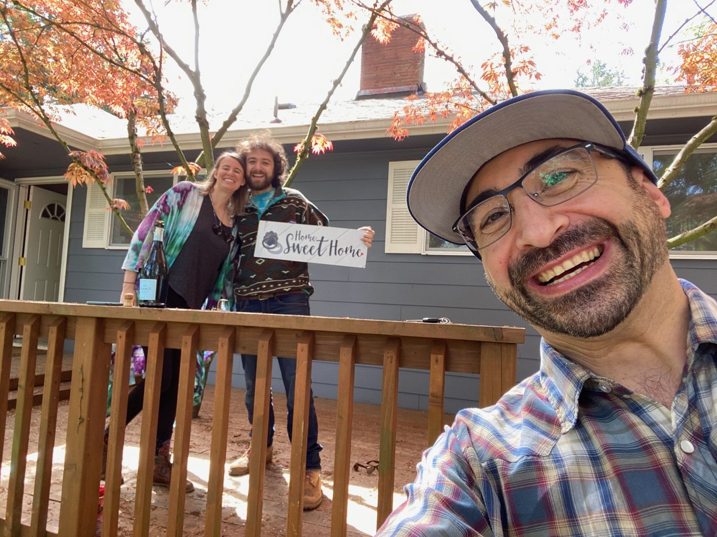 Tim grinning in a close selfie in a bucket hat while a couple hold a Home Sweet Home sign on the back deck of their blue ranch home under a red maple